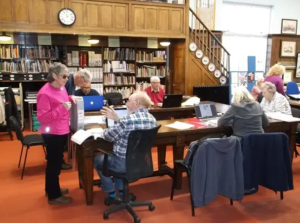 Older adults participate in an activity at tables in a library with bookshelves and wooden panelling.