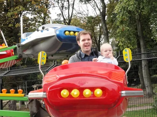 Man and toddler riding in red fairground ride shaped like an aeroplane, with blue plane ride visible behind.