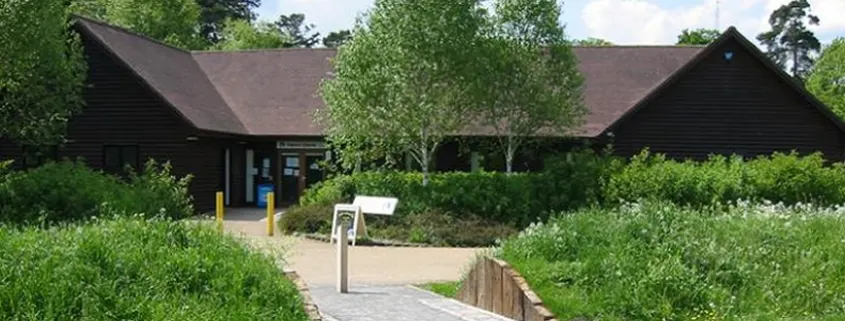 Modern black-clad visitor centre building surrounded by green vegetation and a wooden boardwalk path.