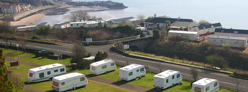 Caravans parked on grassy ground overlooking a coastal bay with beach and clifftop houses.