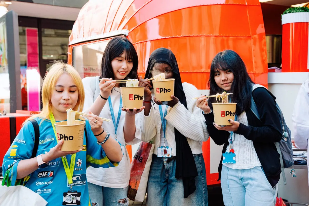Four young people eating pho noodles from branded cups at an outdoor food venue with a large orange umbrella.