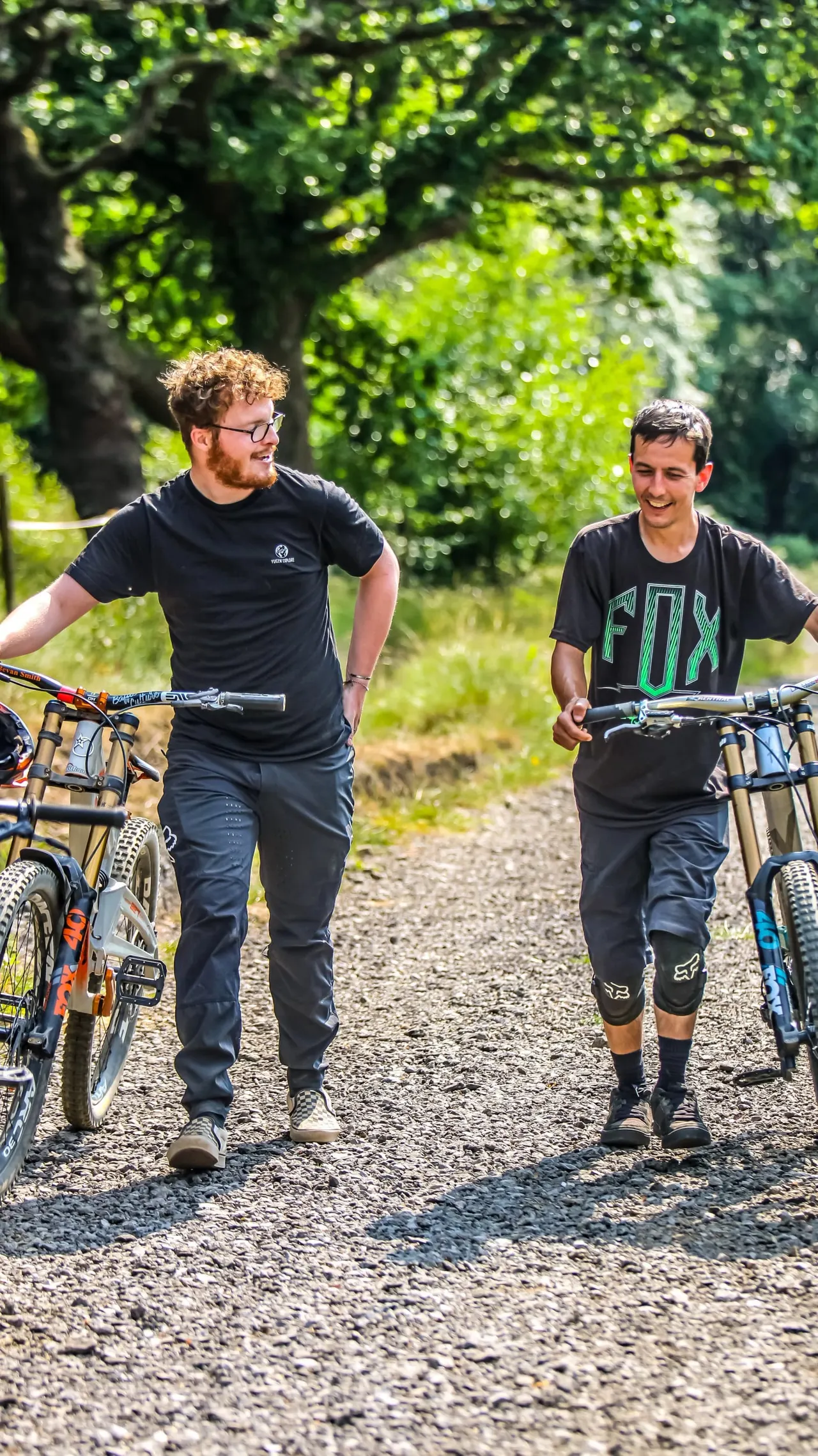 Two young men jogging with bicycles on a gravel path beneath bright green foliage.