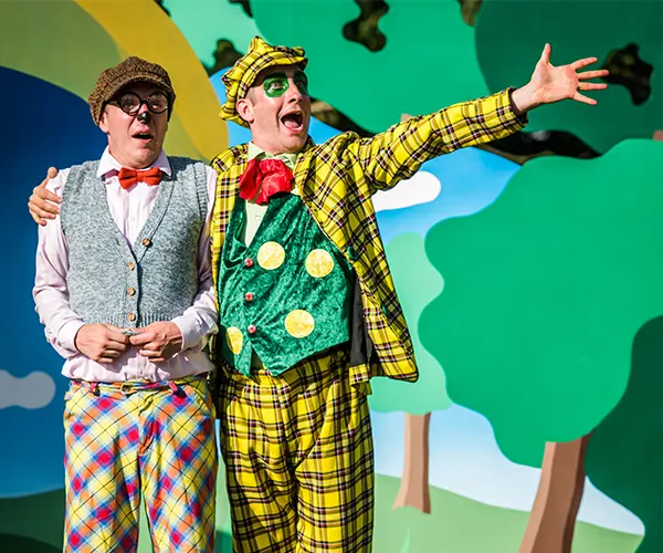 Two performers in colourful clown costumes perform on stage with a painted backdrop of trees and sky.
