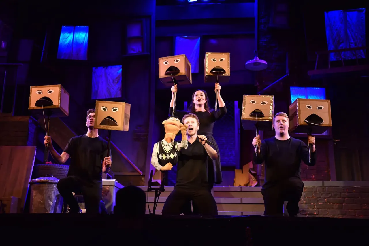 Performers on stage holding large cardboard heads with expressive faces under blue lighting during a theatrical production.