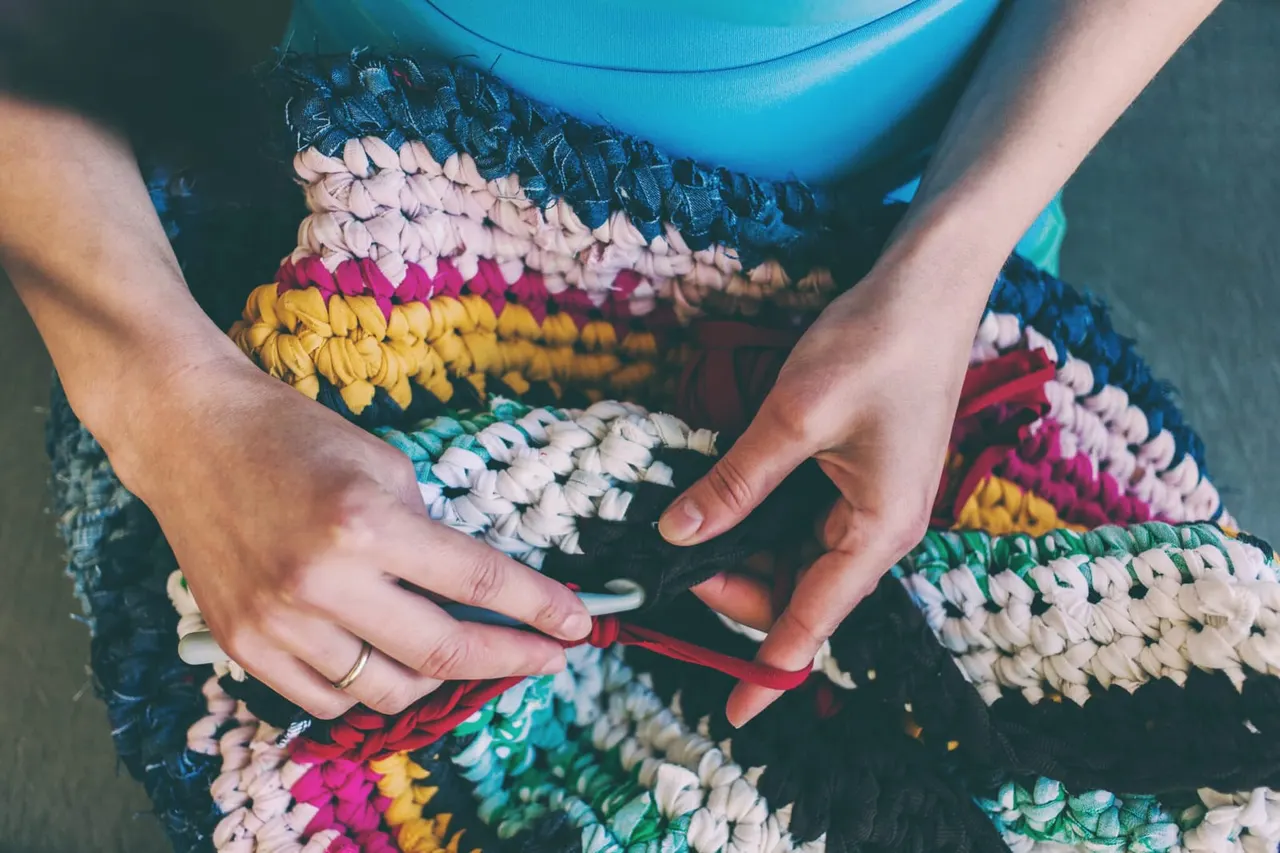 Person's hands holding a colorful woven textile or craft item with blue bucket visible above.