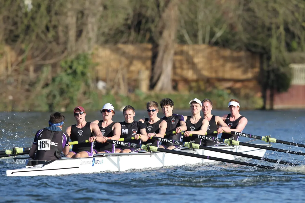 A rowing crew of nine athletes in a racing shell on the water, with trees and buildings visible along the riverbank behind them.