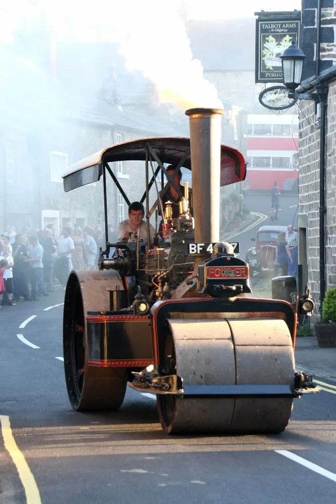 A vintage steam roller with registration BF47 drives down a misty street lined with spectators and period pub signs.