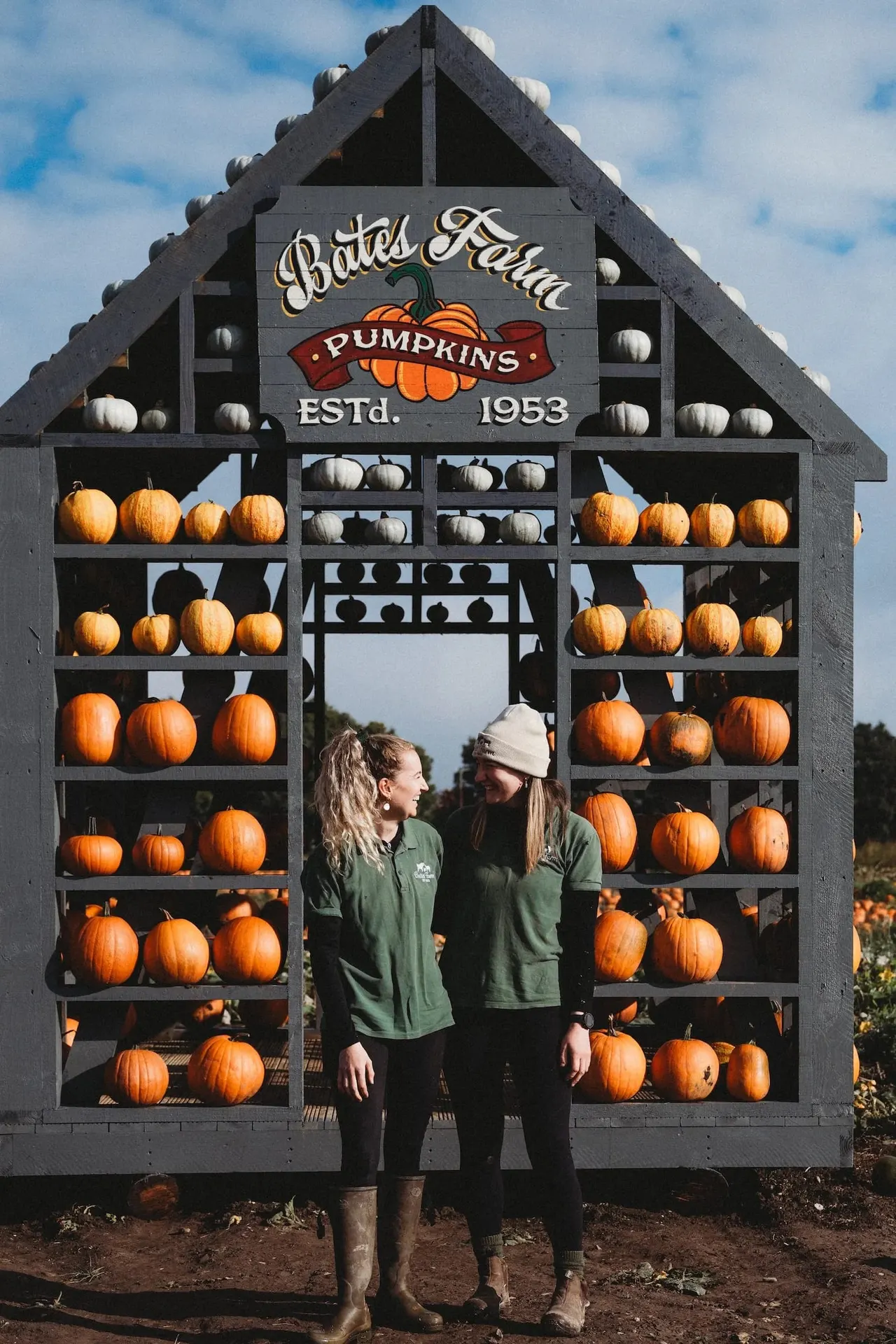 Two visitors stand in front of a large house-shaped pumpkin display stacked with orange and white gourds.