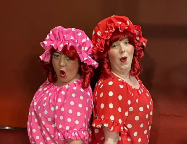 Two performers in matching polka dot costumes and frilly bonnets with styled red curly hair, posing theatrically against a brown backdrop.