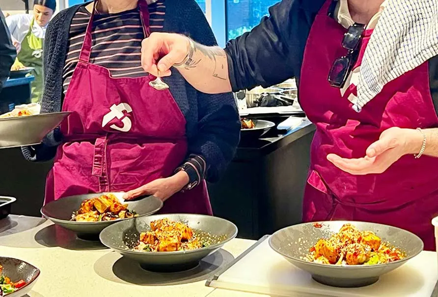 Two people in magenta aprons preparing dishes during a cookery class demonstration.