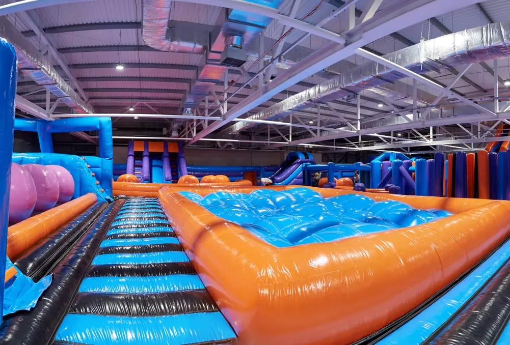 Indoor inflatable play park with blue and orange bounce structures, slides, and obstacle courses under industrial ceiling lights.