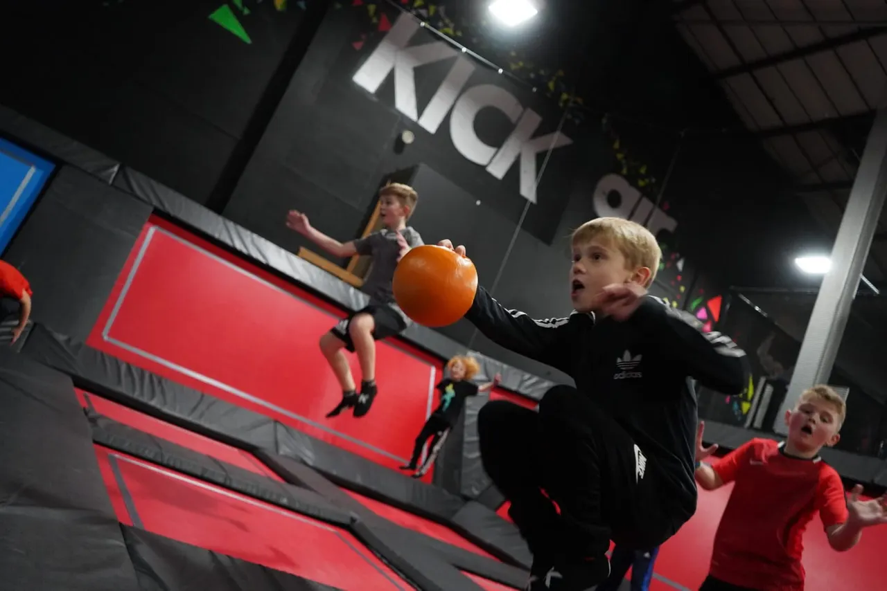 Children playing with a large orange ball inside an indoor trampoline park with red padded surfaces.