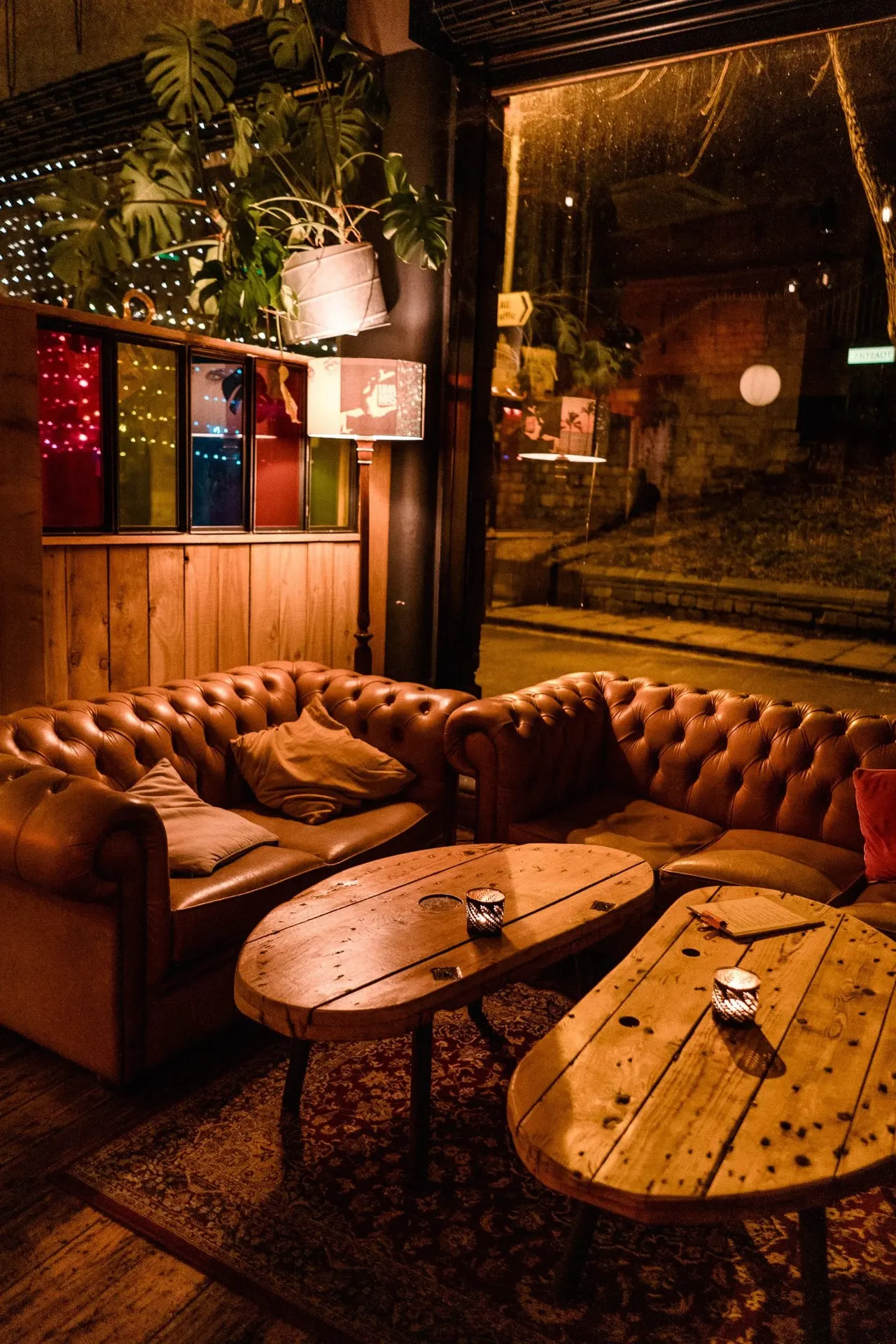 Evening exterior of a leisure venue with brown leather seating, wooden tables and colourful illuminated window panels.