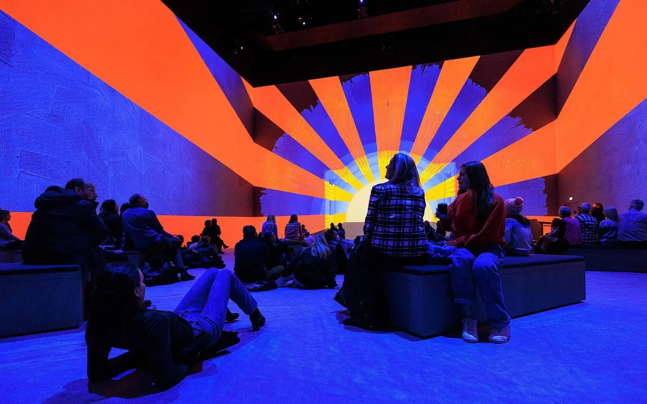 Visitors sit on the floor of an immersive digital art installation bathed in vibrant blue and orange lighting with a radiating sunburst projection.