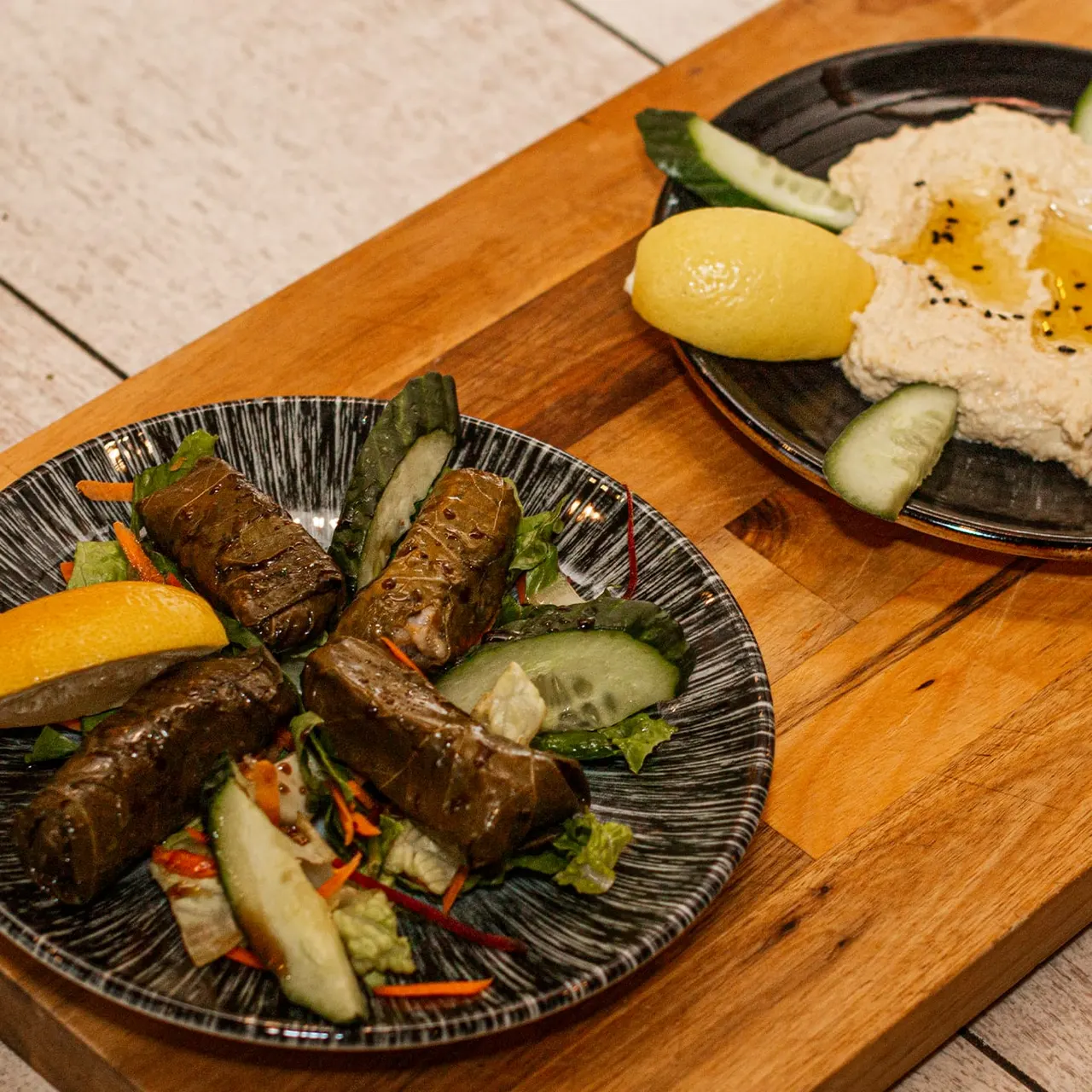 Two plates of Middle Eastern mezze on a wooden board: one with chargrilled meat skewers, cucumber and lemon; another with hummus, cucumber slices and olive oil.