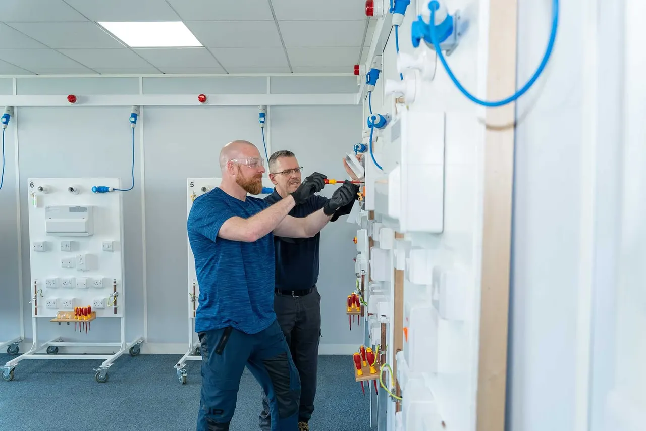 Two men in an electrical training room practice wiring and installation on demonstration boards with cables and components.
