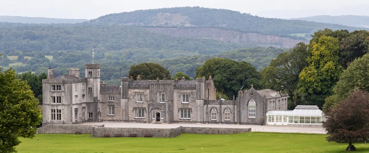 A grand stone castle with attached chapel and modern conservatory sits in expansive grounds, surrounded by woodland and mountains beyond.