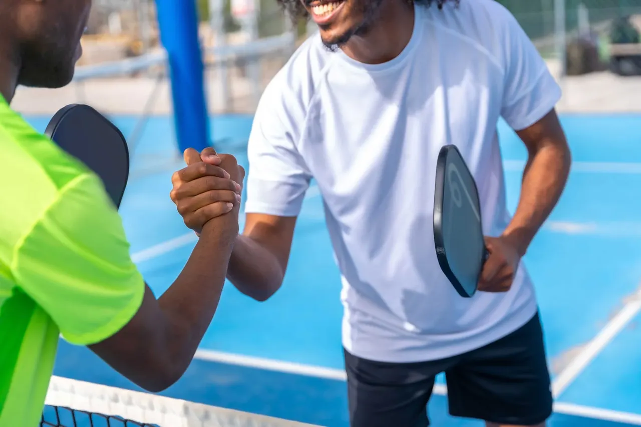 Two people shaking hands on a bright blue pickleball court, both holding paddles.