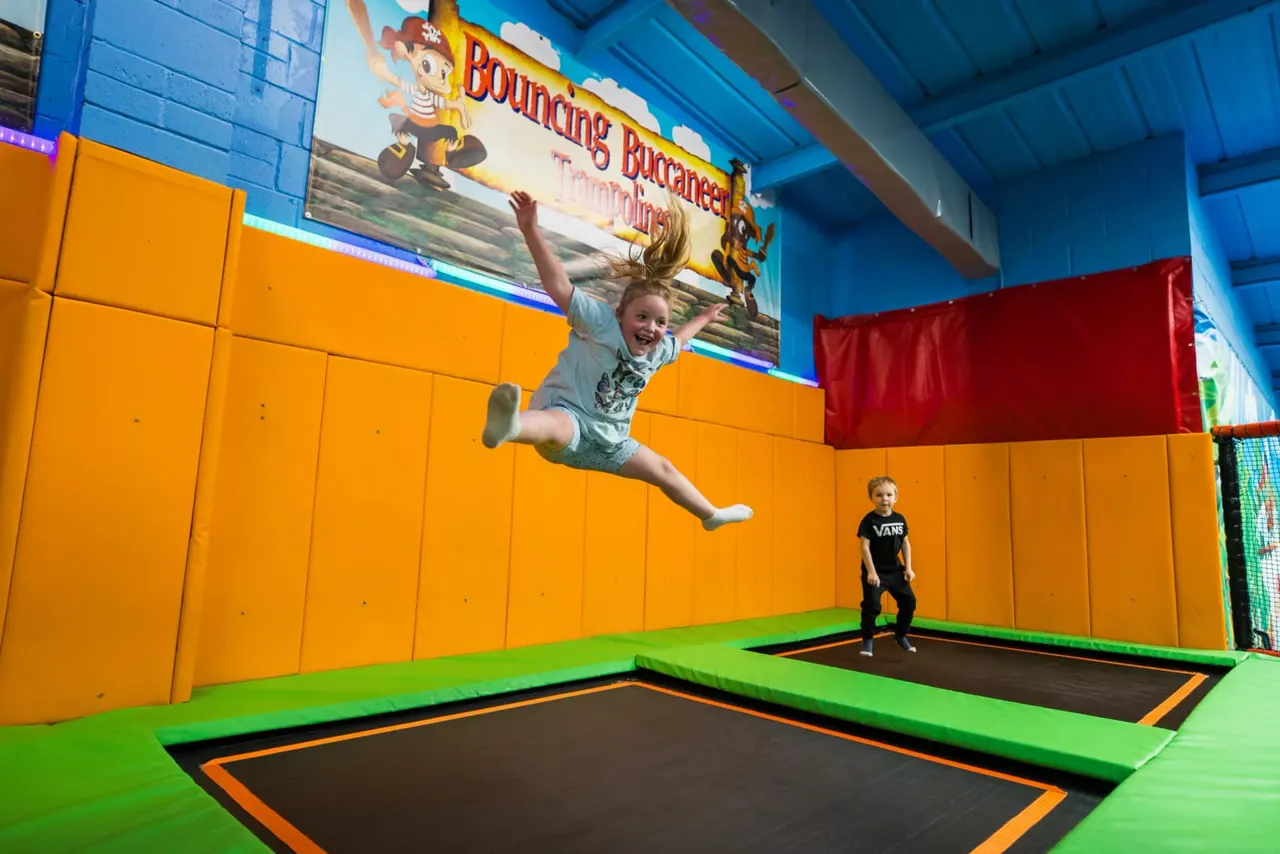 Young child jumping high on a trampoline in a colourful indoor play arena whilst another child watches.