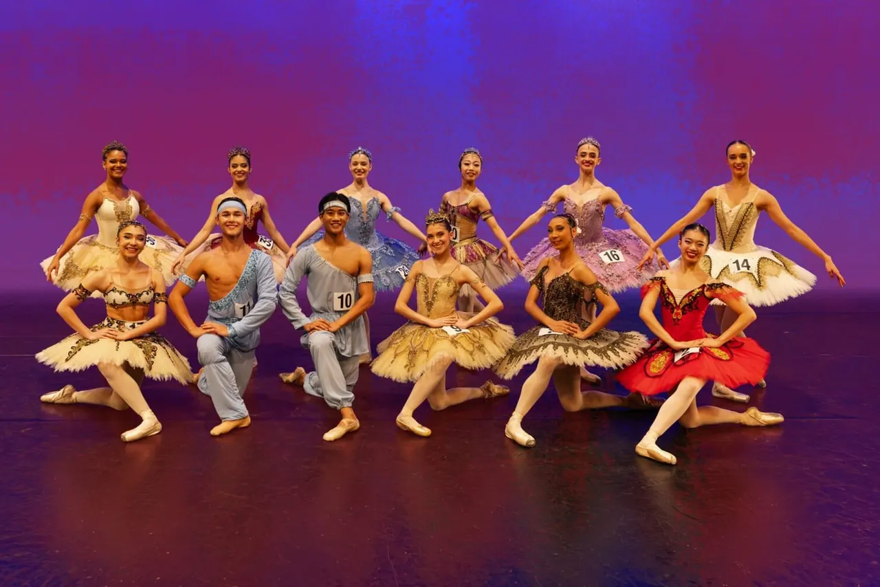 Group of young ballet dancers in costume on stage with purple lighting, wearing tutus and tiaras, numbered for a competition.