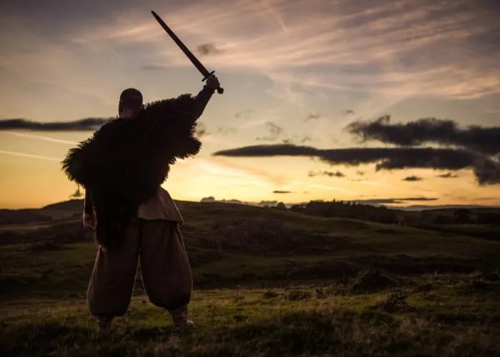 Person in historical costume holding sword aloft in open moorland at sunset.