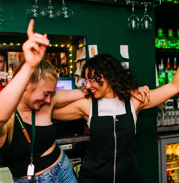 Two women dancing and celebrating together at a lively bar with a green interior and illuminated bottles.