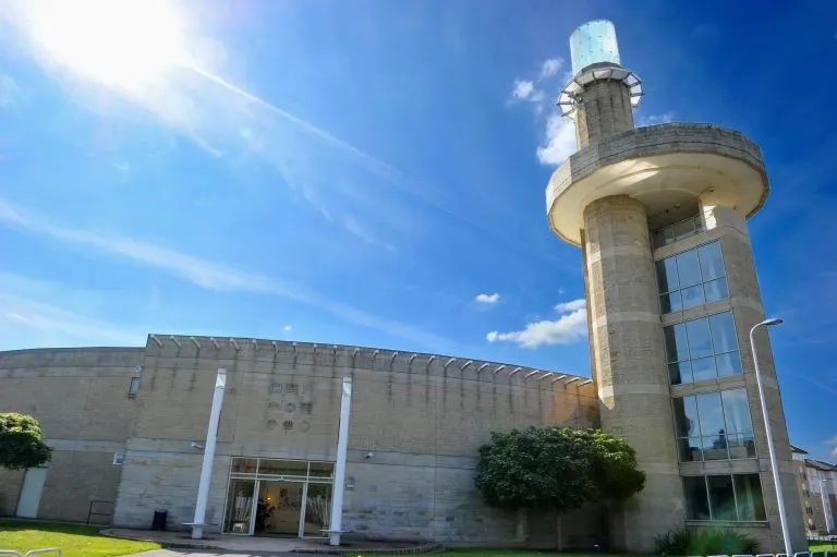 Modernist stone building with a circular tower featuring viewing platform, set against blue sky.