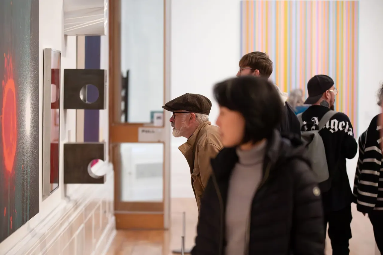 Visitors viewing contemporary artworks in a bright gallery space with colourful striped paintings and geometric sculptures on display.