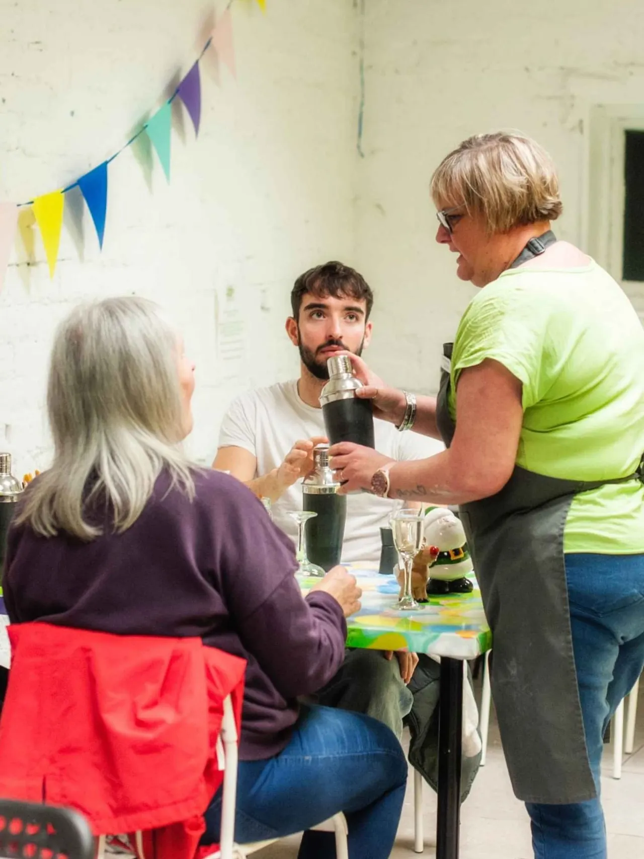 Three people gather around a table at an indoor event, sharing drinks and conversation beneath colourful bunting.