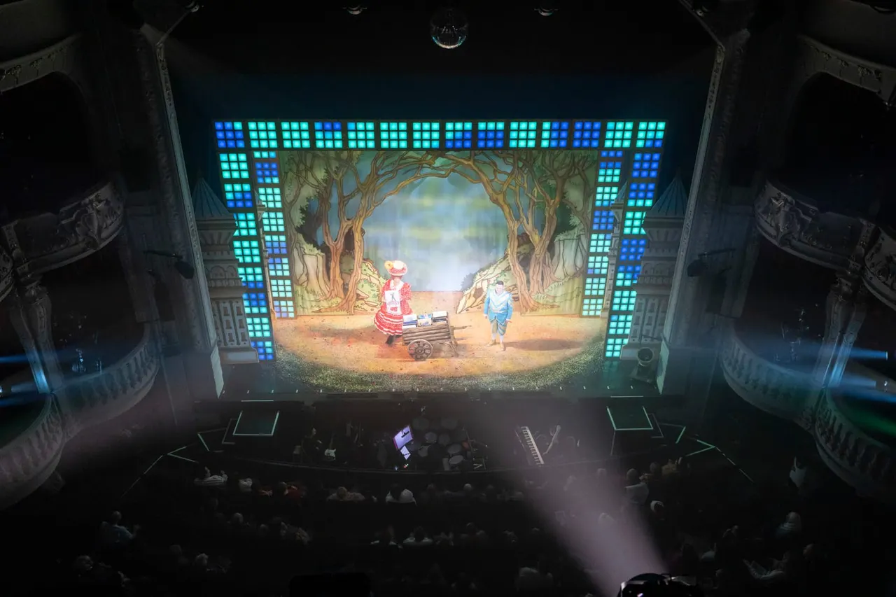 Audience seated in ornate theatre watching animated scene of characters and a cart beneath trees on illuminated stage screen.