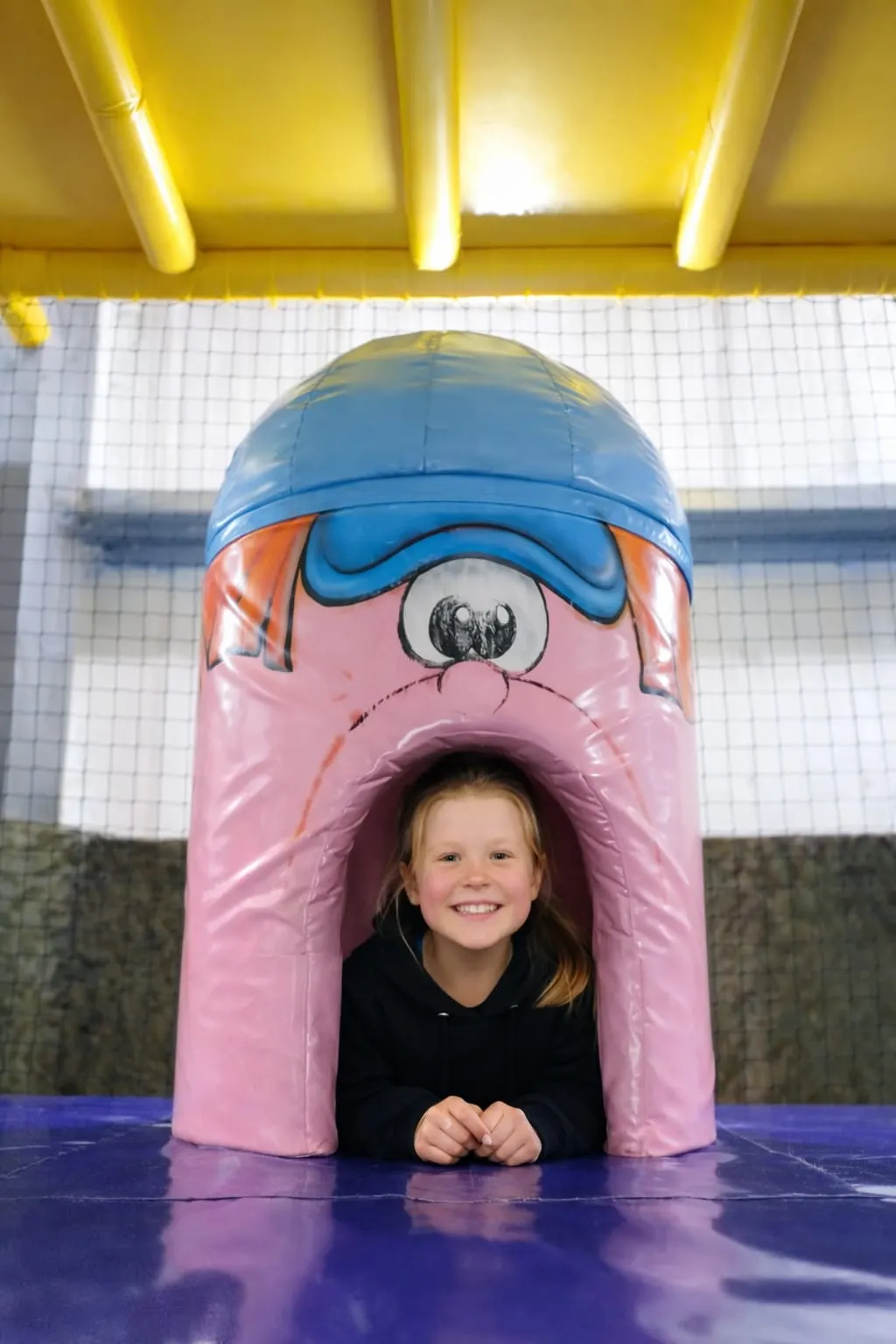 Young child smiling through the entrance of a colourful inflatable play structure indoors.