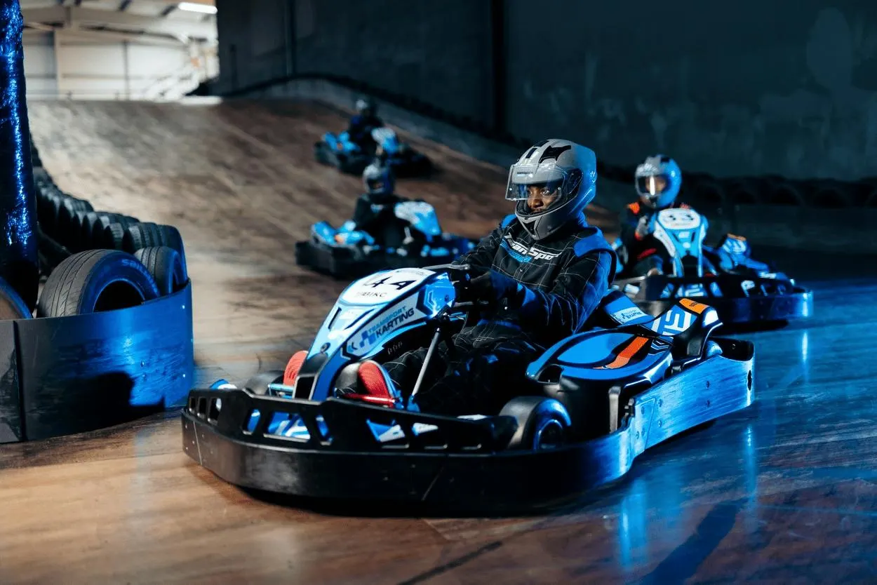 A go-kart racer in blue and black suit and helmet navigates an indoor track past blue barriers, with other karts visible in the background.