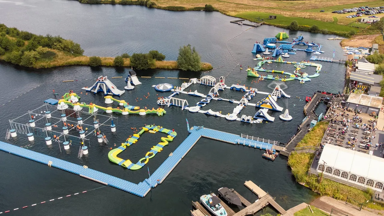 Aerial view of an inflatable water park with multiple obstacle courses and slides on a lake surrounded by green fields and woodland.