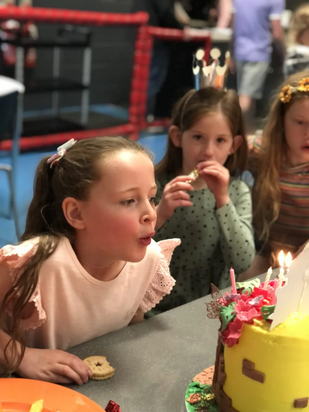 Children at a birthday party blowing out candles on a decorated cake indoors.