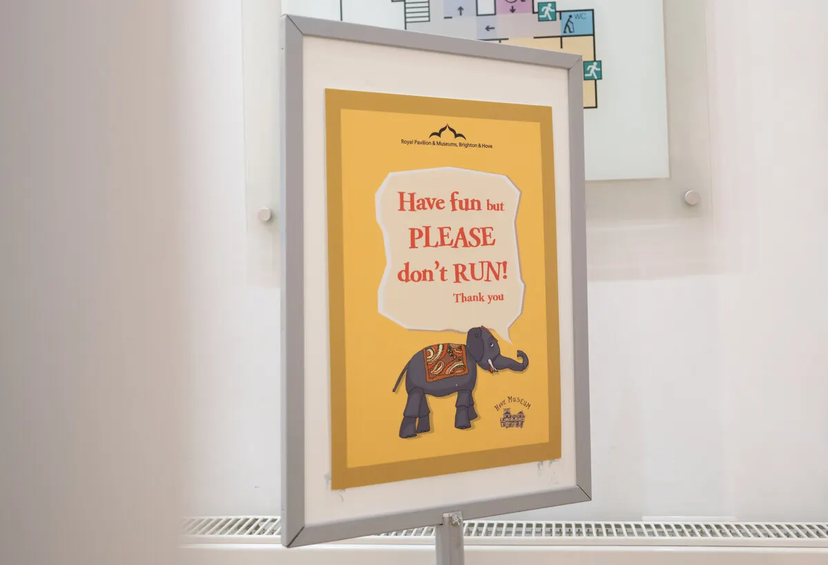 Framed yellow poster with an elephant illustration asking visitors not to run, displayed on a white wall near exit signs.