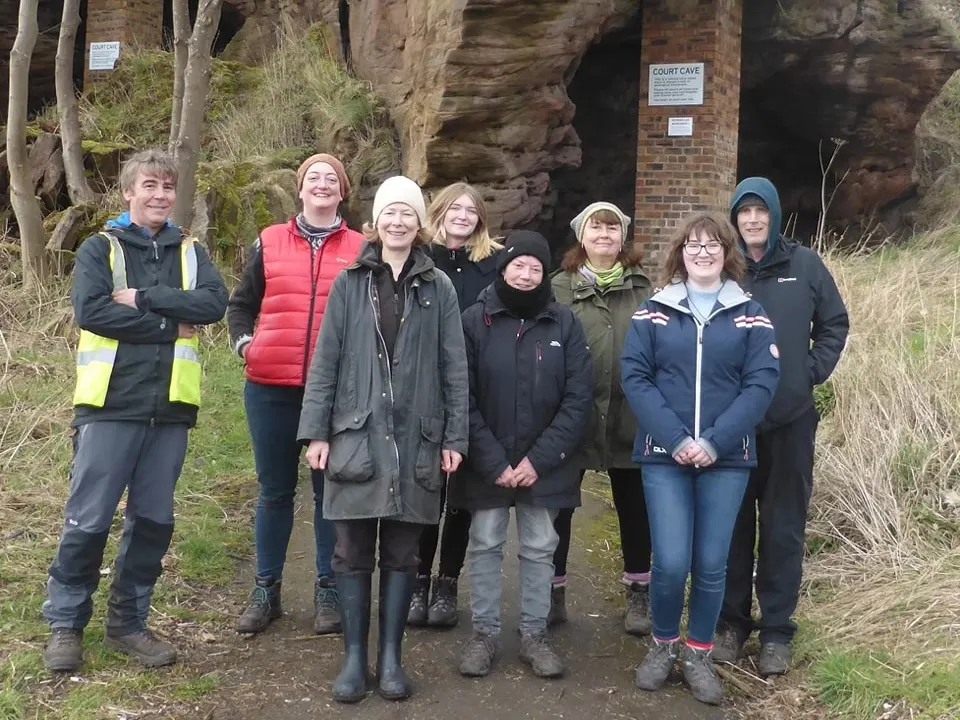 Group of nine visitors standing together outside a cave entrance surrounded by moss-covered rock and bare vegetation.