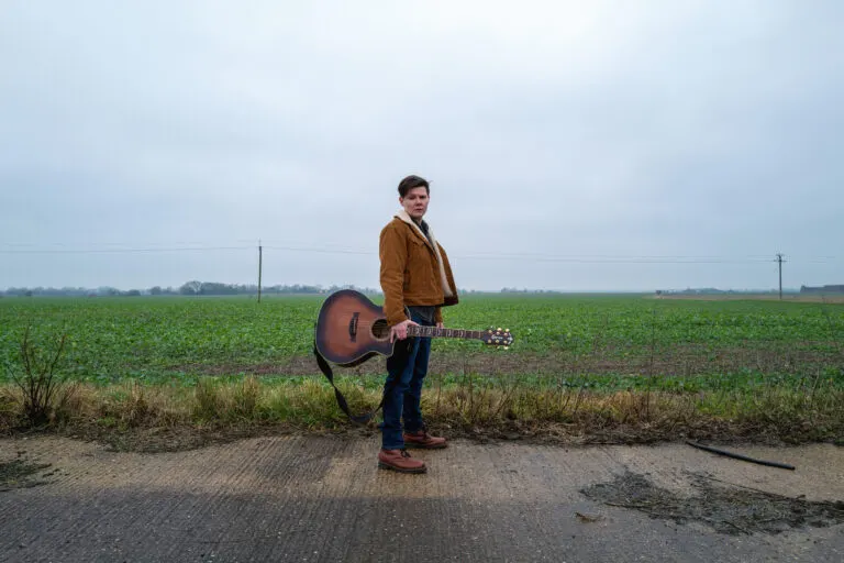 Man in brown jacket holding an acoustic guitar standing on a rural road with flat green fields behind him.