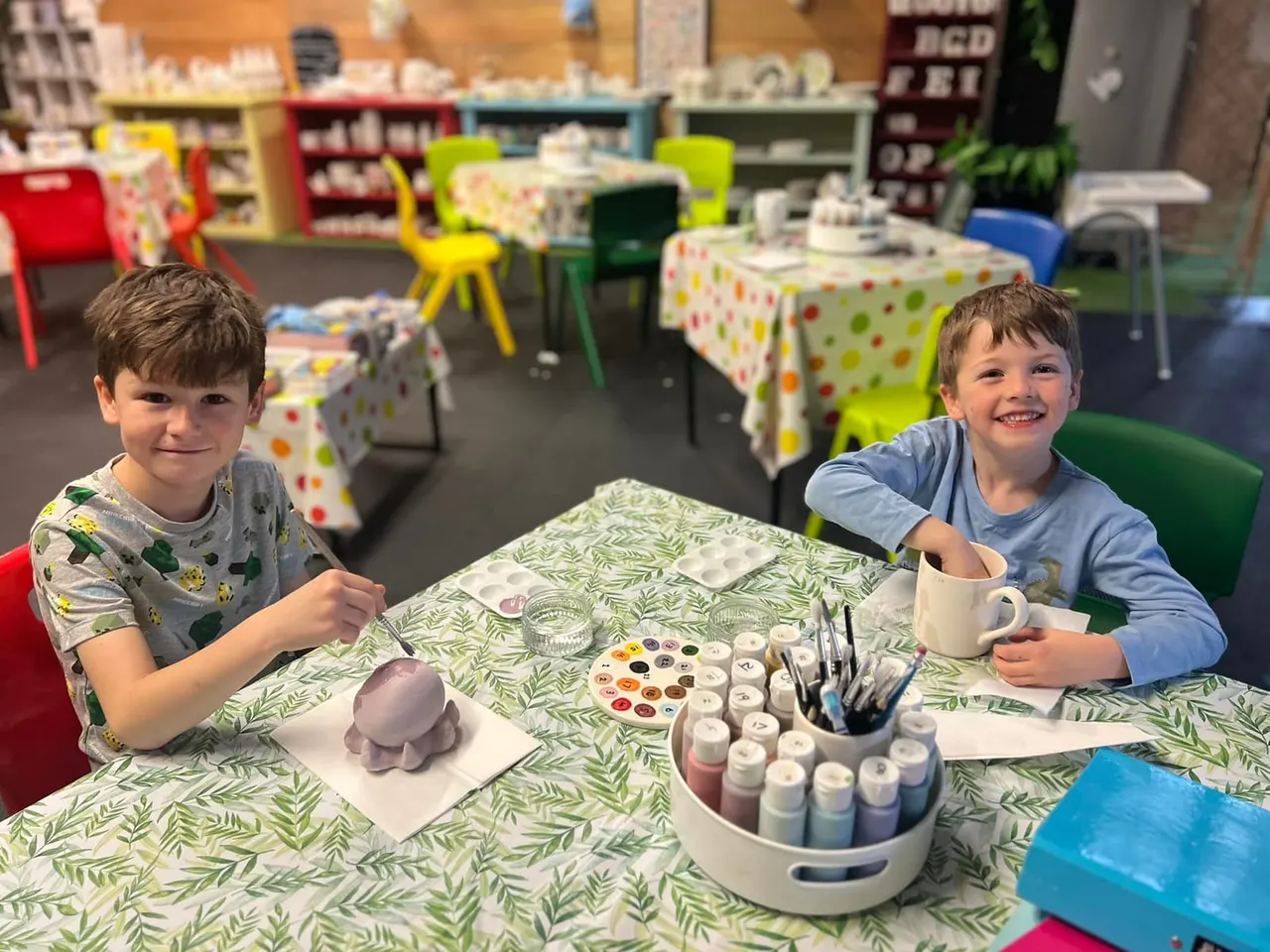 Two young children sitting at a craft table with art supplies, smiling at the camera in a bright activity room.