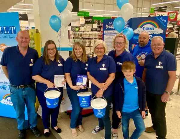 Team of staff and volunteers in matching blue polo shirts posing together in a supermarket with promotional cups and balloons.