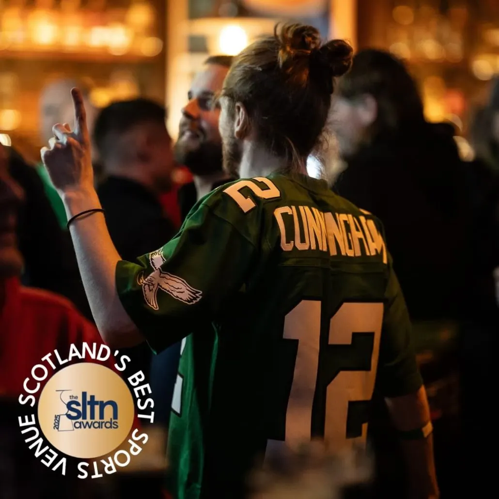 Person in sports jersey celebrating with raised hand among a crowd in an evening venue.