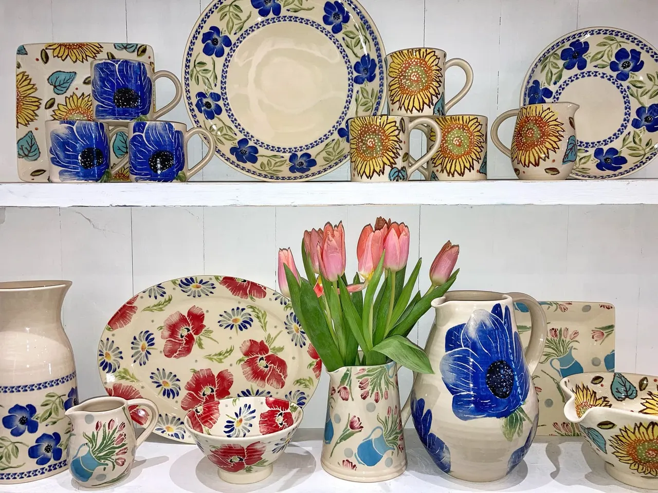 Decorative pottery displayed on white shelves, including hand-painted cups, plates and jugs with floral patterns, and a vase of fresh pink tulips.
