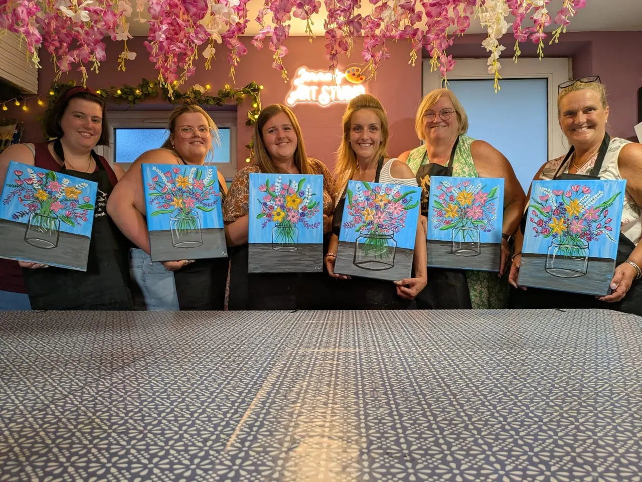 Six women display their matching flower vase paintings indoors beneath hanging pink and yellow flowers.