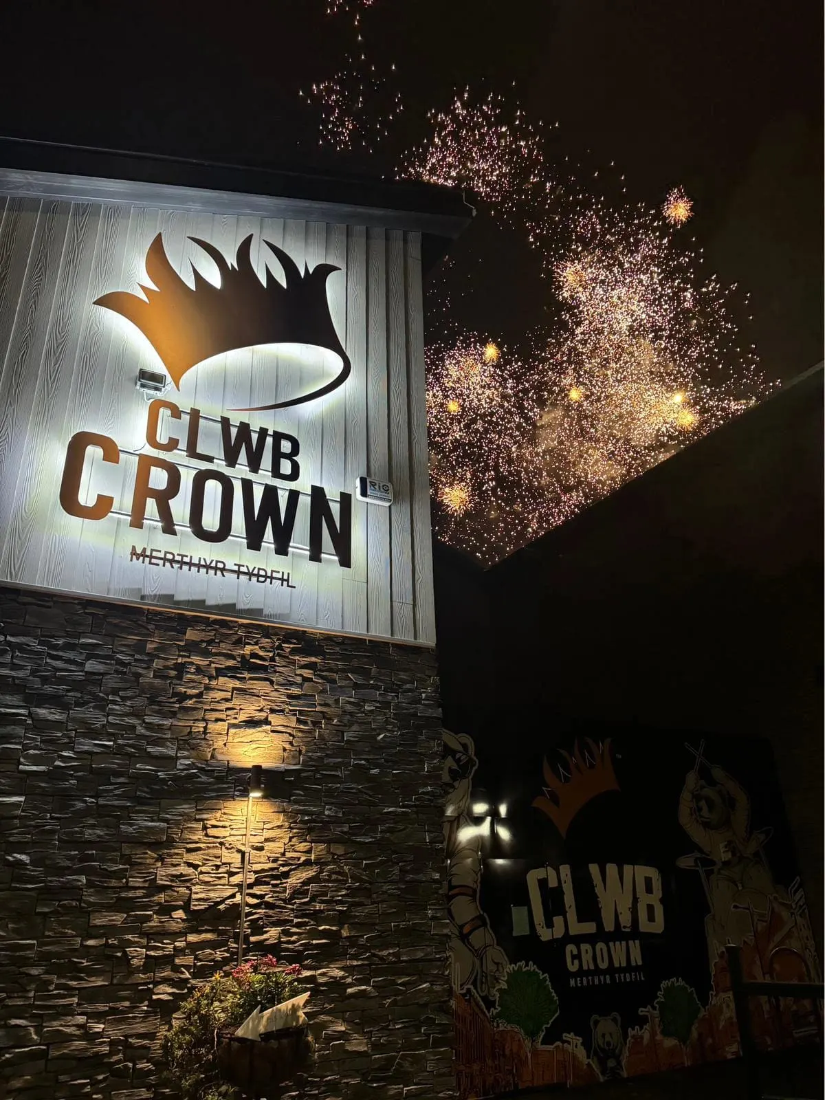 Nighttime scene of an illuminated venue sign for Clwb Crown with fireworks exploding in the dark sky above.