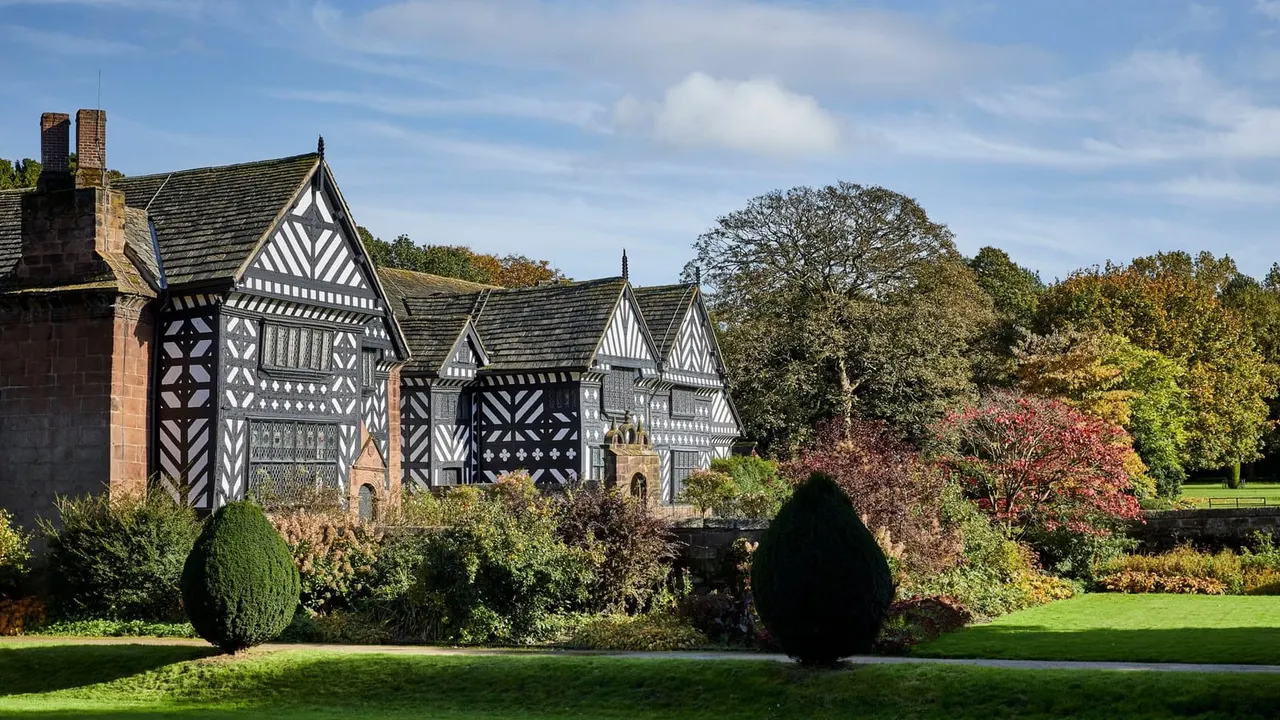 Tudor half-timbered manor house with formal gardens, topiary and autumn trees under blue sky.