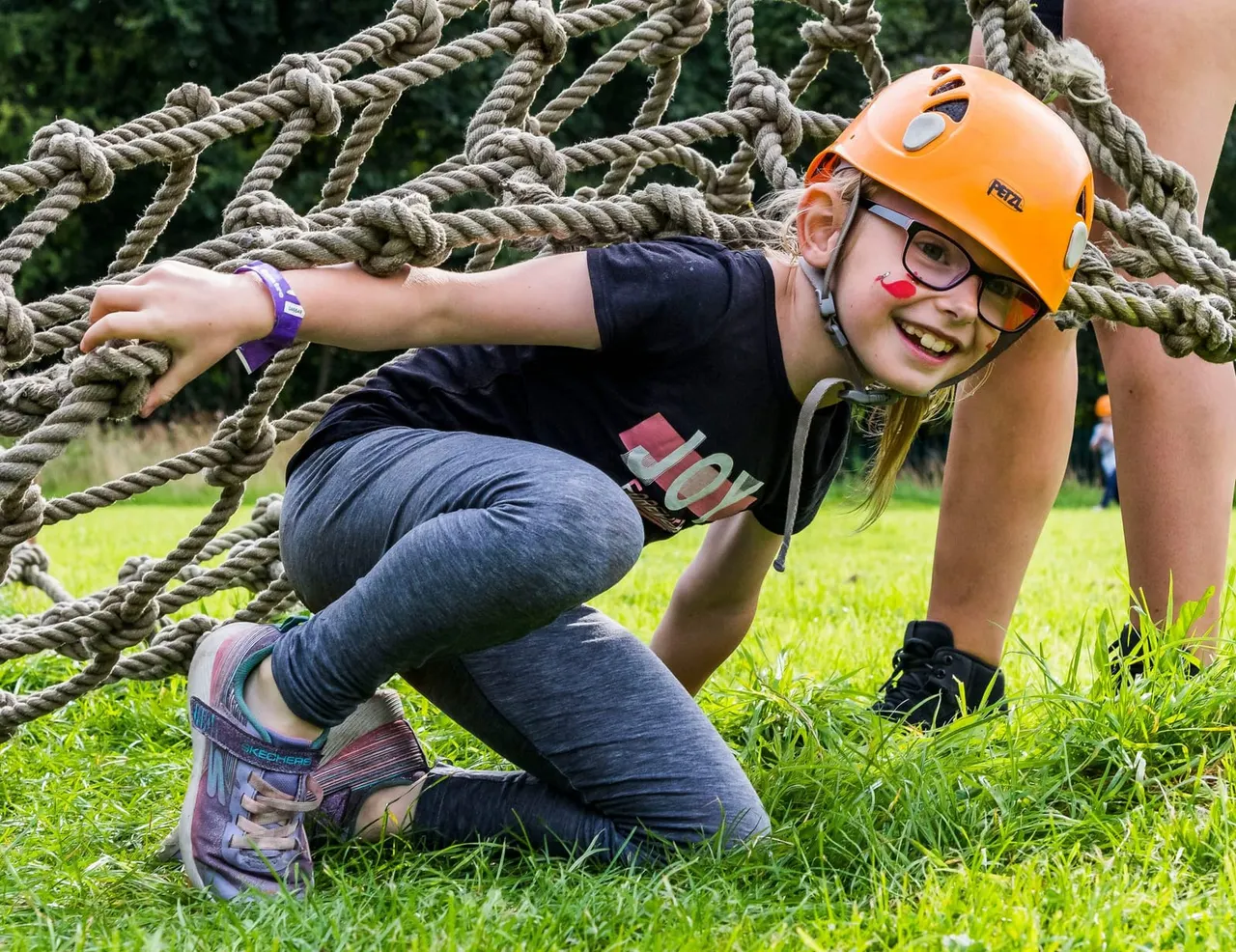 Young person wearing orange safety helmet and harness navigating a rope obstacle course on grass.