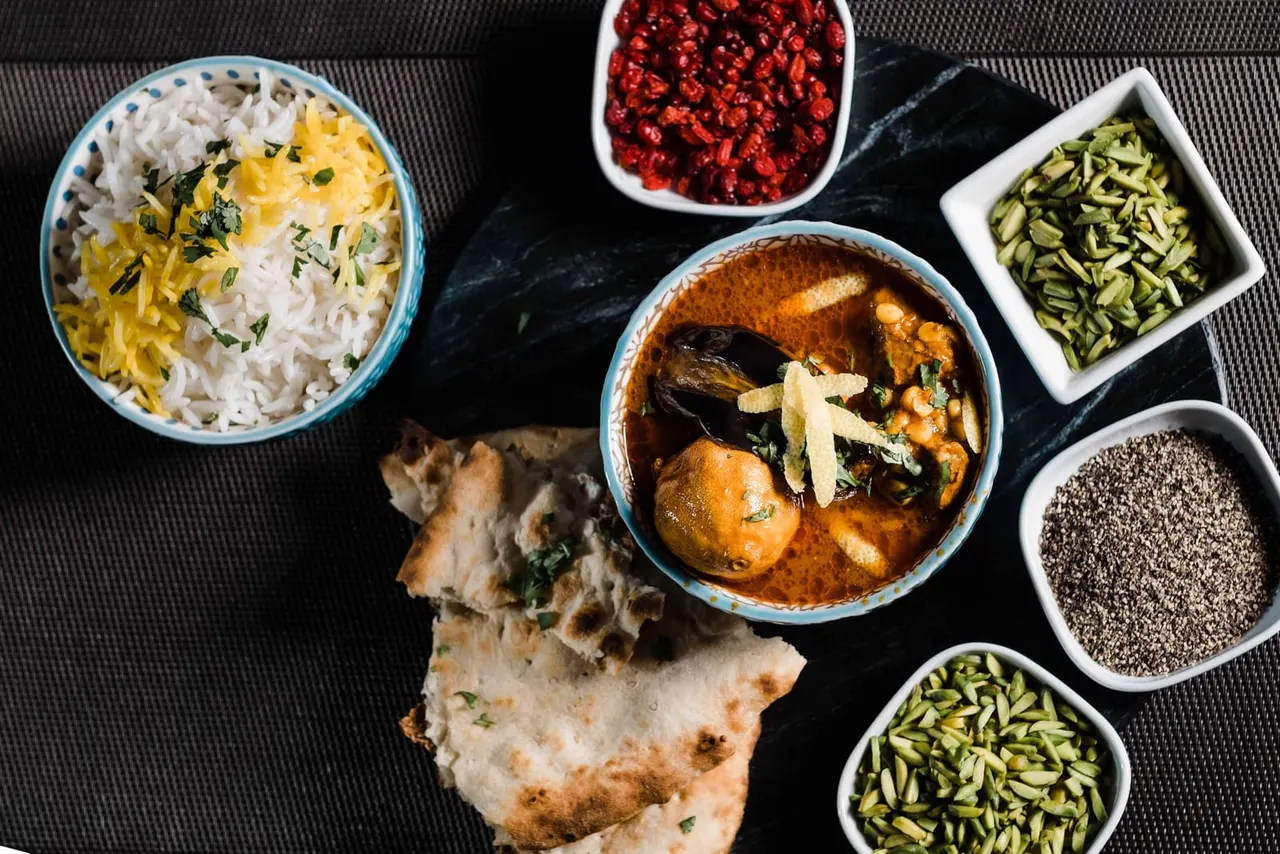 Overhead view of a Persian meal with rice, curry, flatbread, and bowls of seeds, pomegranate, and vegetables on a dark surface.