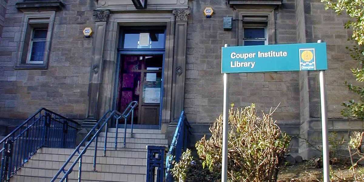 Stone building entrance with purple door, metal handrails on steps, and turquoise Couper Institute Library sign, flanked by autumn foliage.