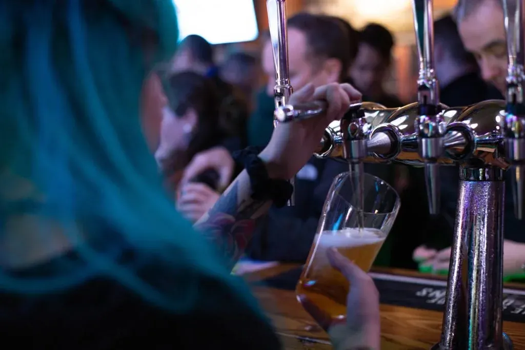 Beer being poured from tap into glass at crowded bar counter with patrons in background.