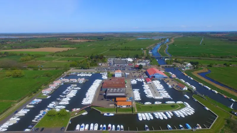 Aerial view of a waterside marina with moored boats, surrounded by green countryside and winding waterways.