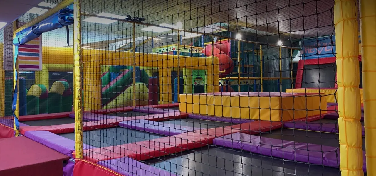 Indoor soft play arena with colourful padded equipment, netting, trampolines and climbing structures for children.